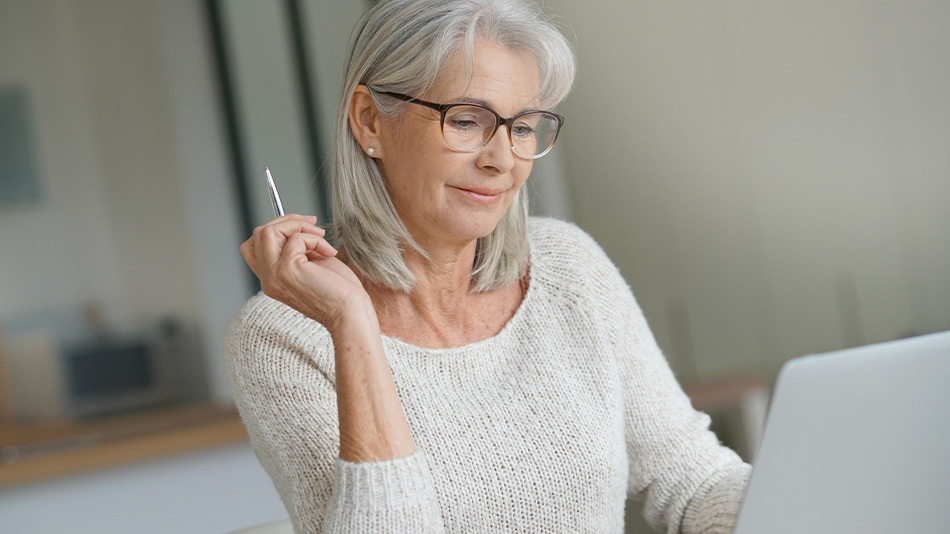 A senior woman with glasses holding a pen while looking at her iPad.
